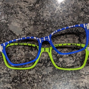 May include: Two pairs of eyeglasses, one blue with white stripes and one green with black stripes, are stacked on top of each other.