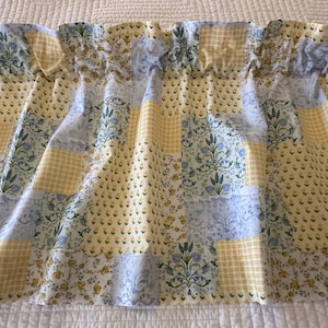 May include: A valance curtain with a patchwork design. The fabric features a mix of patterns, including yellow gingham, floral prints, and blue floral motifs. The curtain is gathered at the top, creating soft folds.