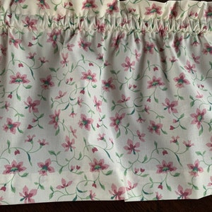 May include: A floral valance featuring a repeating pattern of pink flowers and green leaves on a cream background. The fabric is gathered at the top, creating soft folds. This window treatment adds a touch of vintage charm.