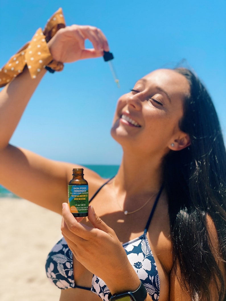 K&ouml;nnte beinhalten: Eine Frau h&auml;lt eine Flasche Skin Food Organics Vitamin C Serum. Die Flasche ist blau und gelb mit wei&szlig;em Text. Die Frau tr&auml;gt ein schwarz-wei&szlig;es Blumen-Bikini-Oberteil und eine goldene Halskette.