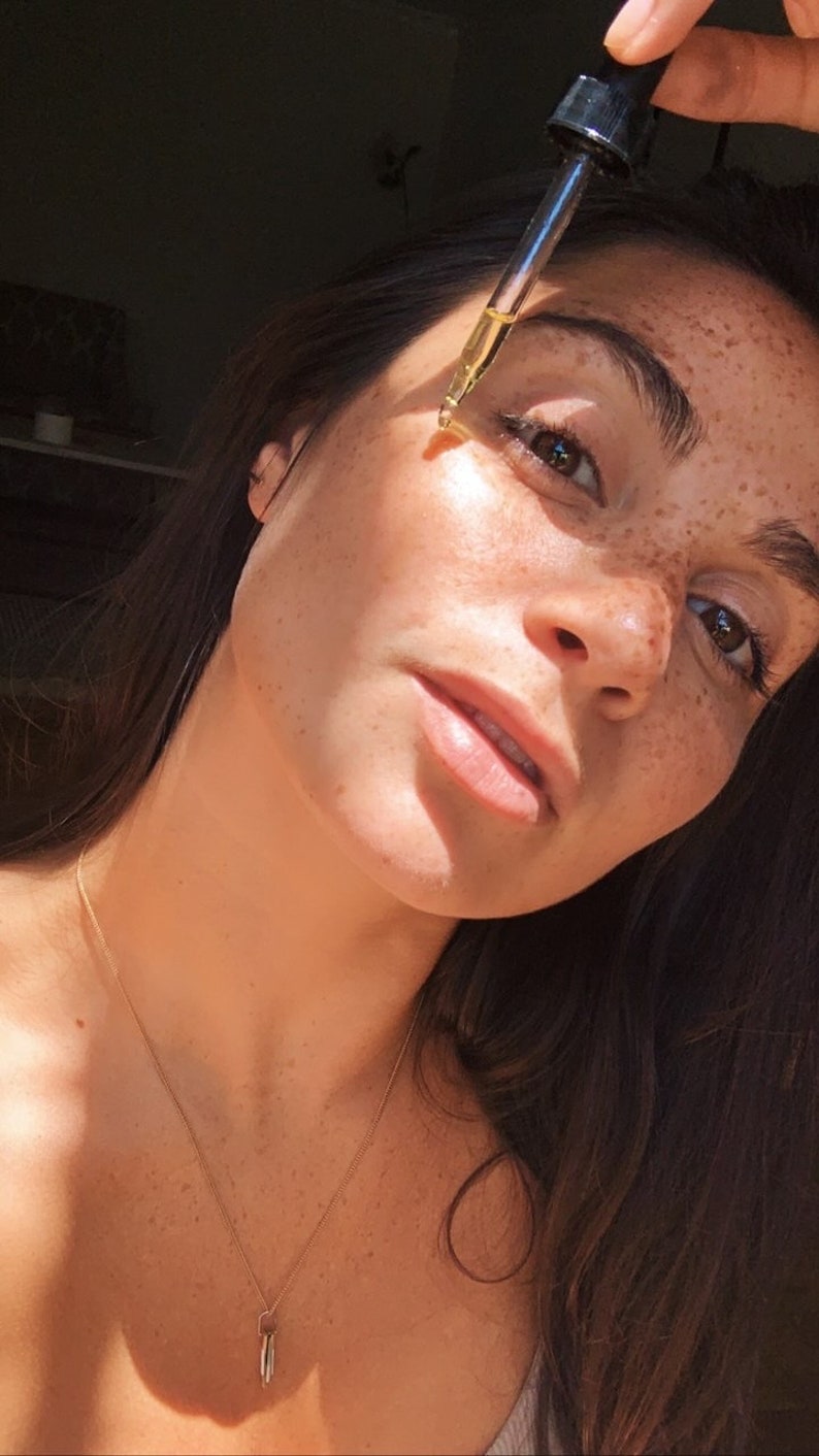 K&ouml;nnte beinhalten: Eine Frau mit Sommersprossen tr&auml;gt ein klares Gesichts&ouml;l mit einer Pipette auf. Sie tr&auml;gt eine goldene Halskette mit einem kleinen Anh&auml;nger.