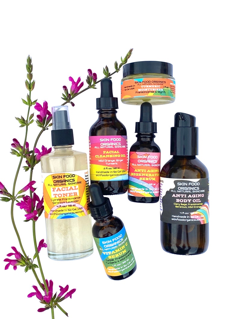 K&ouml;nnte beinhalten: Ein Set aus sechs bernsteinfarbenen Glasflaschen mit Tropfern und Etiketten f&uuml;r Gesichtswasser, Reinigungs&ouml;l, Anti-Aging-Serum, Vitamin-C-Serum und Anti-Aging-K&ouml;rper&ouml;l. Die Flaschen sind auf einer wei&szlig;en Oberfl&auml;che mit einem Zweig lila Bl&uuml;ten angeordnet.