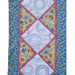 May include: A blue and green table runner with a diamond pattern of white fabric with embroidered circles. The runner has a pink border.