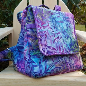 Peut inclure: Un sac à dos en tissu à motif violet et bleu avec une fermeture à rabat. Le sac à dos a une seule sangle et est fait d'un tissu doux et matelassé.