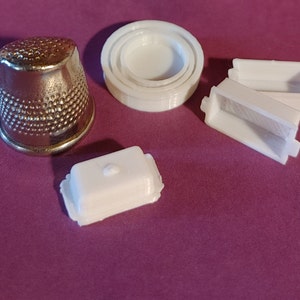 May include: A silver thimble with a dimpled surface is next to several white plastic items. These include nested circular containers and rectangular containers, possibly for crafts or small items.