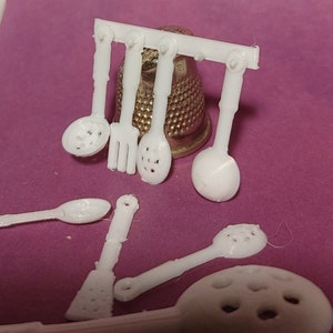 Puede incluir: Un juego de utensilios de cocina de plástico blanco, que incluye una cuchara ranurada, un tenedor y una cuchara, colgados de un estante. También hay tres cucharas adicionales en la superficie rosa.