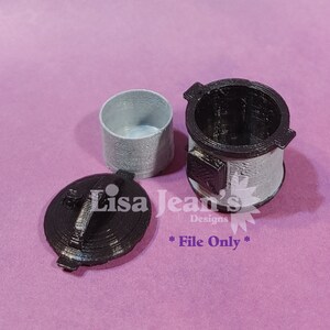 May include: A miniature 3D printed stove with a black lid and a grey body. The stove has a small door and a chimney. The stove is sitting on a pink surface.