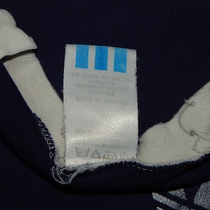 May include: A white tag with blue stripes and text that reads "Care instructions, 100% cotton, wash with similar colors, do not bleach, do not tumble dry, iron at low temperature, do not dry clean, the brand with the 3 stripes, made in the USA".
