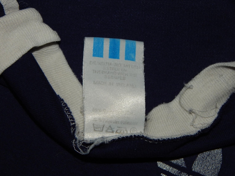 May include: A white tag with blue stripes and text that reads "Care instructions, 100% cotton, wash with similar colors, do not bleach, do not tumble dry, iron at low temperature, do not dry clean, the brand with the 3 stripes, made in the USA".