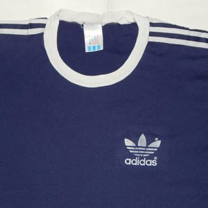 May include: A navy blue t-shirt with white stripes on the shoulders and a white ribbed collar. The shirt has a silver Adidas logo with the word "adidas" underneath.