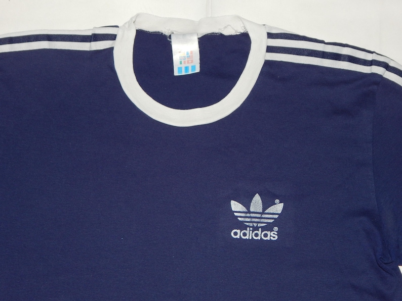 May include: A navy blue t-shirt with white stripes on the shoulders and a white ribbed collar. The shirt has a silver Adidas logo with the word "adidas" underneath.
