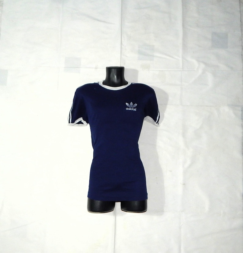 May include: A navy blue t-shirt with white trim around the neck and sleeves. The shirt has a white Adidas logo with the word "adidas" underneath.