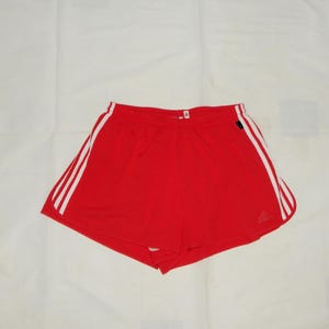 May include: Red athletic shorts with white stripes down the sides. The shorts have a drawstring waist and a small logo on the left leg.