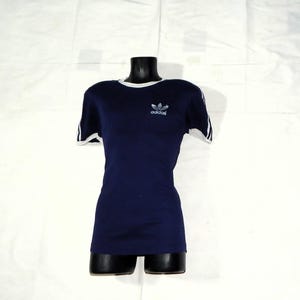 May include: A navy blue t-shirt with white trim around the neck and sleeves. The shirt has a white Adidas logo on the front.