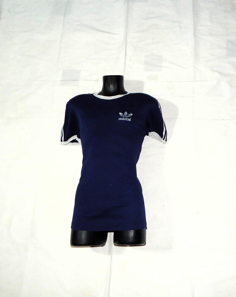 May include: A navy blue t-shirt with white trim around the neck and sleeves. The shirt has a white Adidas logo on the front.