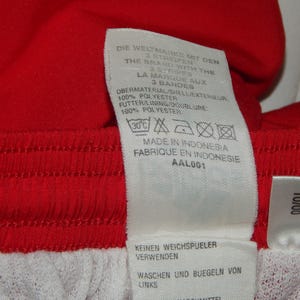 May include: A white tag with black text and symbols. The tag is attached to a red and white garment. The text on the tag reads: "DIE WELTMARKE MIT DEN 3 BANDES THE BRAND WITH THE 3 BANDES LA MARQUE AUX 3 BANDES OBERMATERIAL/SIELLEKTENEUPC 100% POLYESTER FUTTERLINING DOUBLURE 100% POLYESTER MADE IN INDONESIA FABRIQUE EN INDONESIE AAL001 KEINEN WEICHSPJELER VERWENDEN WASCHEN UND BUEGELN VON LINKS 10/00"