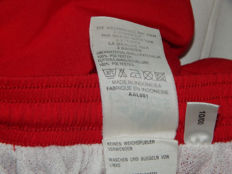 May include: A white tag with black text and symbols. The tag is attached to a red and white garment. The text on the tag reads: "DIE WELTMARKE MIT DEN 3 BANDES THE BRAND WITH THE 3 BANDES LA MARQUE AUX 3 BANDES OBERMATERIAL/SIELLEKTENEUPC 100% POLYESTER FUTTERLINING DOUBLURE 100% POLYESTER MADE IN INDONESIA FABRIQUE EN INDONESIE AAL001 KEINEN WEICHSPJELER VERWENDEN WASCHEN UND BUEGELN VON LINKS 10/00"