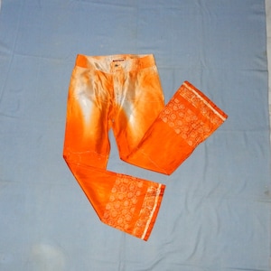 Può includere: Pantaloni arancioni con un motivo bianco e arancione. I pantaloni hanno una gamba larga e un bordo bianco intorno al fondo.