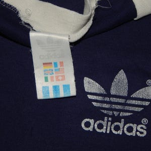 May include: A navy blue t-shirt with a white Adidas logo and a white tag with the Adidas logo and a series of flags.