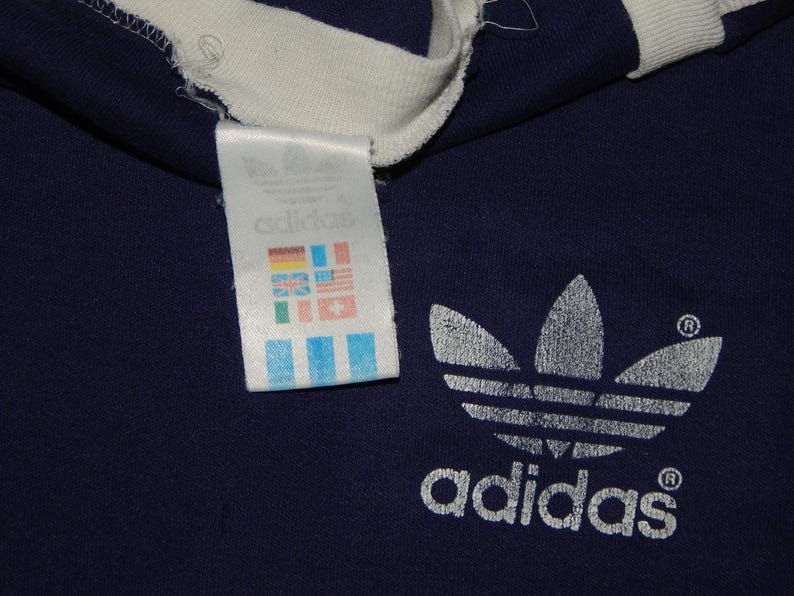 May include: A navy blue t-shirt with a white Adidas logo and a white tag with the Adidas logo and a series of flags.