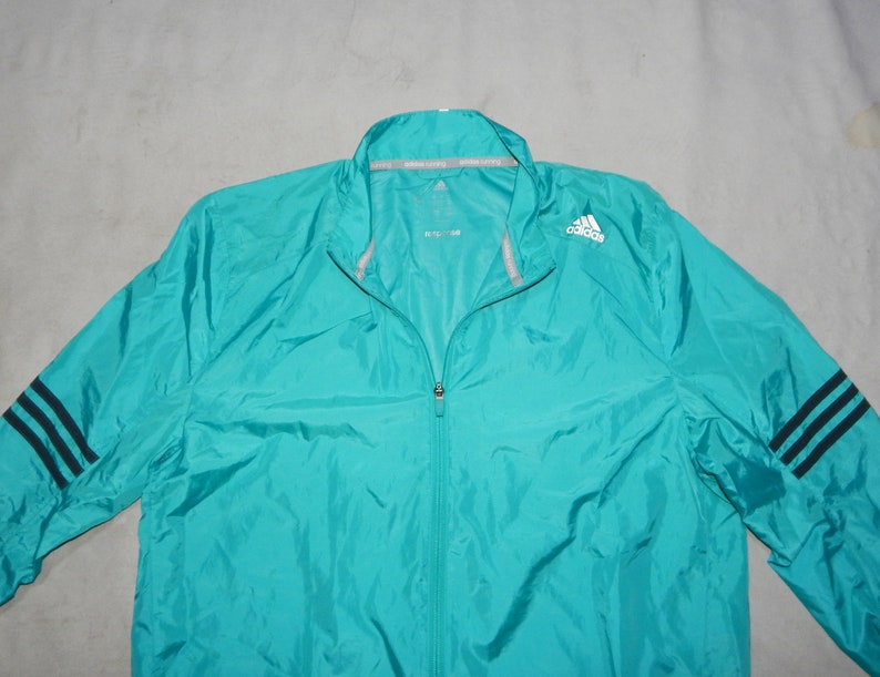 May include: A turquoise blue lightweight jacket with a zipper closure and black and white stripes on the sleeves. The jacket has the Adidas logo on the right chest.