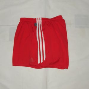 May include: Red athletic shorts with white stripes down the sides. The shorts have a black Adidas logo on the left leg.