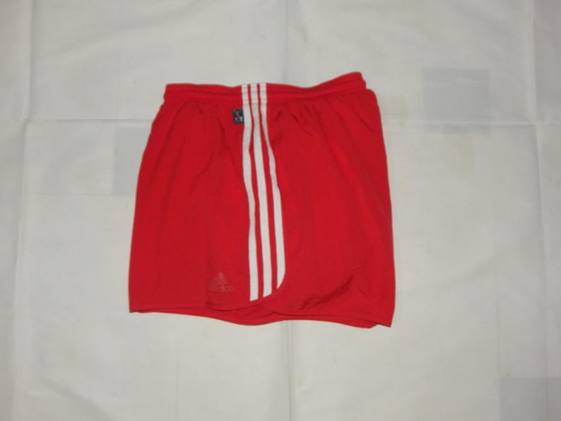May include: Red athletic shorts with white stripes down the sides. The shorts have a black Adidas logo on the left leg.