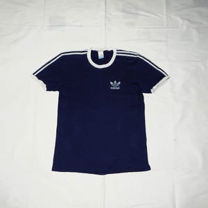 May include: A navy blue t-shirt with white trim around the neck and sleeves. The shirt has a white Adidas logo with three stripes on the front.