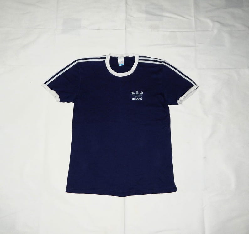 May include: A navy blue t-shirt with white trim around the neck and sleeves. The shirt has a white Adidas logo with three stripes on the front.