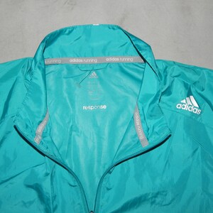 May include: A teal blue windbreaker jacket with a zipper closure and a white Adidas logo on the right side. The jacket has a gray Adidas running label on the inside of the collar.