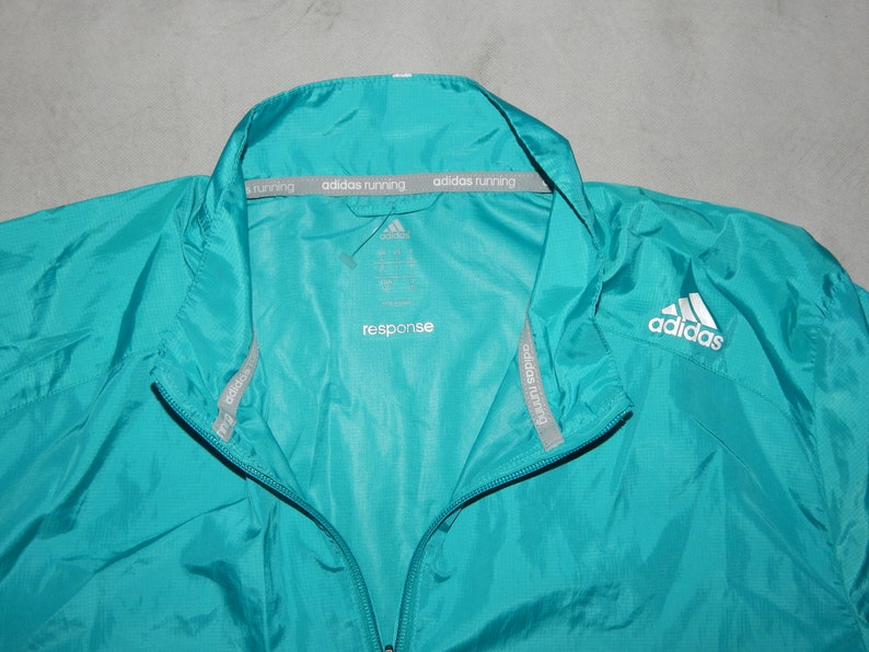May include: A teal blue windbreaker jacket with a zipper closure and a white Adidas logo on the right side. The jacket has a gray Adidas running label on the inside of the collar.