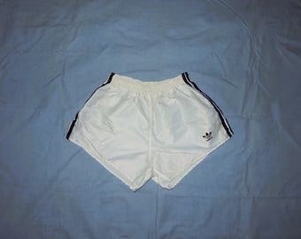 May include: White athletic shorts with navy blue stripes down the sides and a small logo. The shorts are made of a shiny, lightweight material, likely nylon or a similar fabric. The shorts are likely vintage.