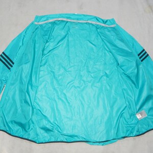 May include: A turquoise blue windbreaker jacket with black stripes on the sleeves. The jacket is unzipped and laid flat on a white surface.
