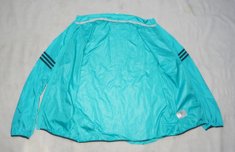 May include: A turquoise blue windbreaker jacket with black stripes on the sleeves. The jacket is unzipped and laid flat on a white surface.