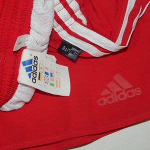 May include: Red athletic shorts with white stripes and the Adidas logo. The shorts have a white waistband and a tag that says "ClimaLite" and "Adidas" with country flags.