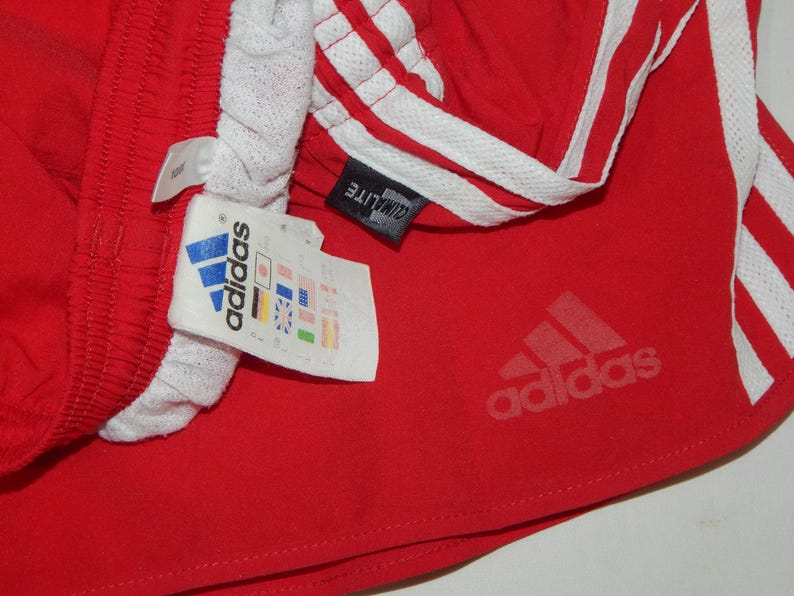 May include: Red athletic shorts with white stripes and the Adidas logo. The shorts have a white waistband and a tag that says "ClimaLite" and "Adidas" with country flags.