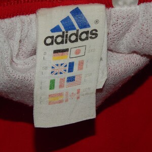 May include: A white tag with the Adidas logo and text, along with a row of small flags representing different countries. The tag is attached to a red and white garment.