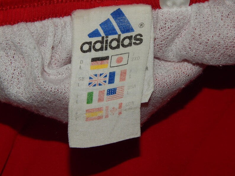 May include: A white tag with the Adidas logo and text, along with a row of small flags representing different countries. The tag is attached to a red and white garment.