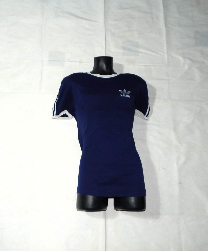 May include: A navy blue t-shirt with white trim around the neck and sleeves. The shirt has a white Adidas logo with the word "adidas" underneath.