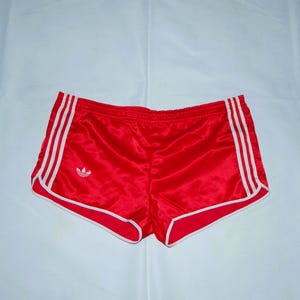 May include: Red athletic shorts with white trim and three white stripes down the sides. The shorts feature the Adidas trefoil logo on the left leg. The shorts are made of a shiny, satin-like material.
