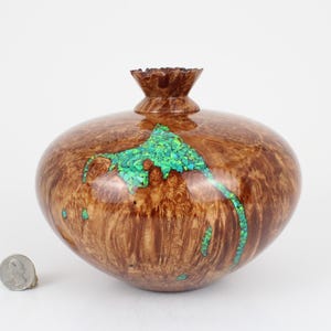 May include: A polished wooden vessel with a rounded body and a flared rim, featuring a prominent natural wood grain pattern and an inlaid section of bright green crushed stone. A silver-colored US quarter coin is placed next to the vessel.