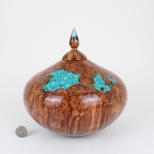 May include: A polished wooden vessel with a rounded body and a pointed lid, featuring inlaid turquoise-colored stone fragments. The wood has a swirling grain pattern. A small circular metal coin is placed next to the vessel for scale.