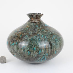 May include: A rounded wooden vessel with a polished finish, featuring dark brown and grey swirling wood grain patterns inlaid with bright turquoise fragments. The vessel has a narrow, flared neck and is shown next to a US quarter coin for scale.