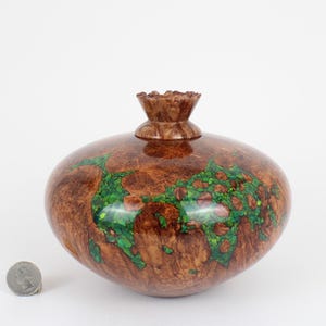 May include: A polished, rounded wooden vessel with a natural brown burl grain pattern and inlaid bright green crushed stone accents. The vessel features a small, flared neck and is displayed next to a silver US quarter for scale reference.