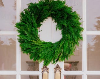 Op de afbeelding: Een cirkelvormige krans gemaakt van dicht, levendig groen groenblijvend loof. De krans heeft een volle, weelderige opstelling van naaldachtige bladeren, waardoor een uniforme en ronde vorm ontstaat die op een witte deur wordt getoond.