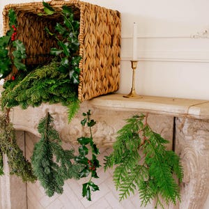 May include: A rectangular woven wicker basket filled with green pine branches and holly sprigs with red berries, placed on a stone mantel. Five individual evergreen and holly branches are suspended from the mantel edge by thin twine.