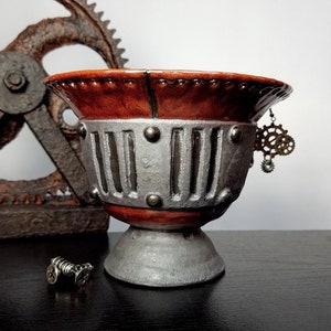 Pode incluir: Uma tigela de cerâmica com acabamento prateado e marrom, com um design steampunk com engrenagens e rebites. A tigela tem uma borda larga e uma base redonda.