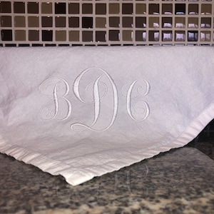 May include: White kitchen towel with a monogram of the letters "BDC" embroidered in silver thread.