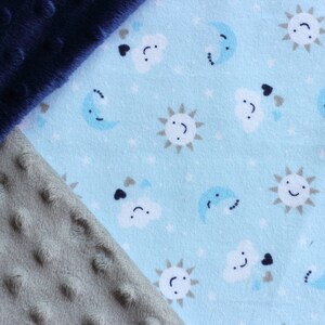 May include: A close-up of two fabric swatches. One is a dark blue plush fabric with small raised dots. The other is a light gray plush fabric with small raised dots. The second fabric is layered over the first fabric and is partially visible. The second fabric has a light blue background with a repeating pattern of white clouds, suns, and moons.