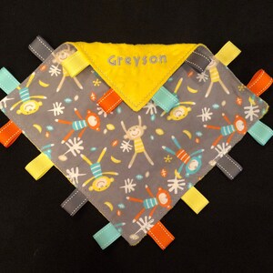 May include: A gray and yellow baby security blanket with a monkey print and the name Greyson embroidered on the yellow fabric. The blanket has multiple colorful ribbons attached to the edges.